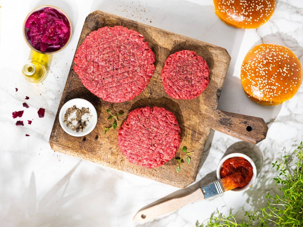 To rå hamburgerkjøttdeiger på en skjærebrett med to hamburgerbrød og ulike tilbehør rundt, som ketchup, sennep og krydder.