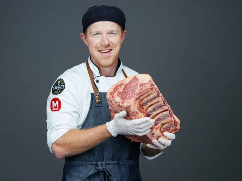 Person som holder et stort stykke rått kjøtt i hendene, iført hvit kokkejakke og forkle, mot en grå bakgrunn.