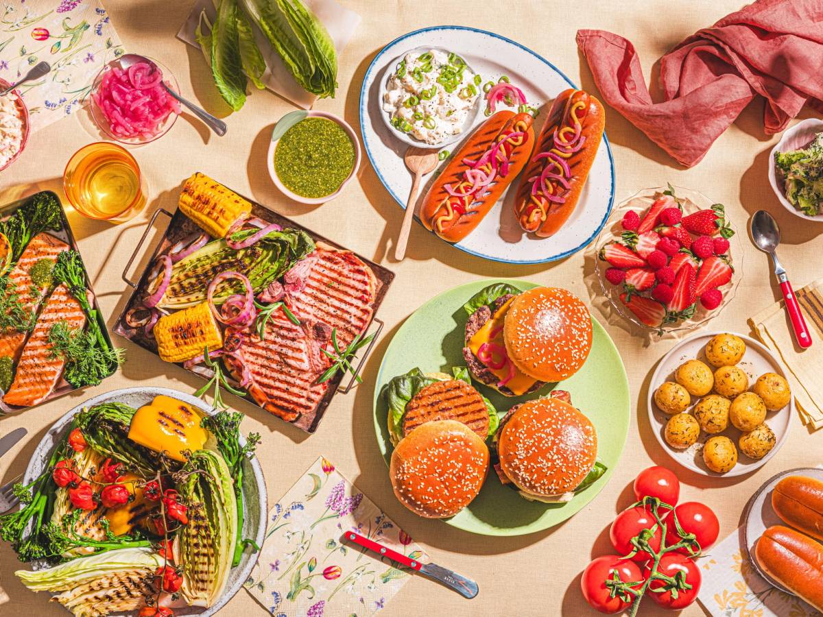 Et bord dekket med ulike sommermatretter, inkludert burgere, pølser, salater og frukt, med en rosa klut og bestikk rundt.