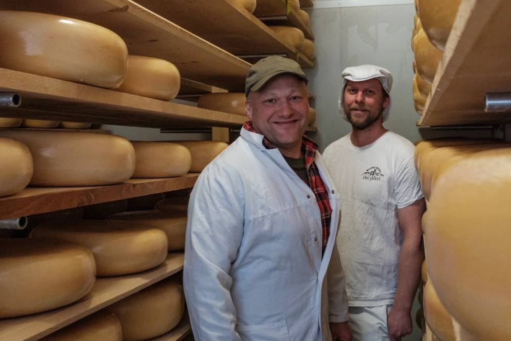 To personer står mellom hyller med ost i en ostebutikk eller lager, begge iført hvite klær.