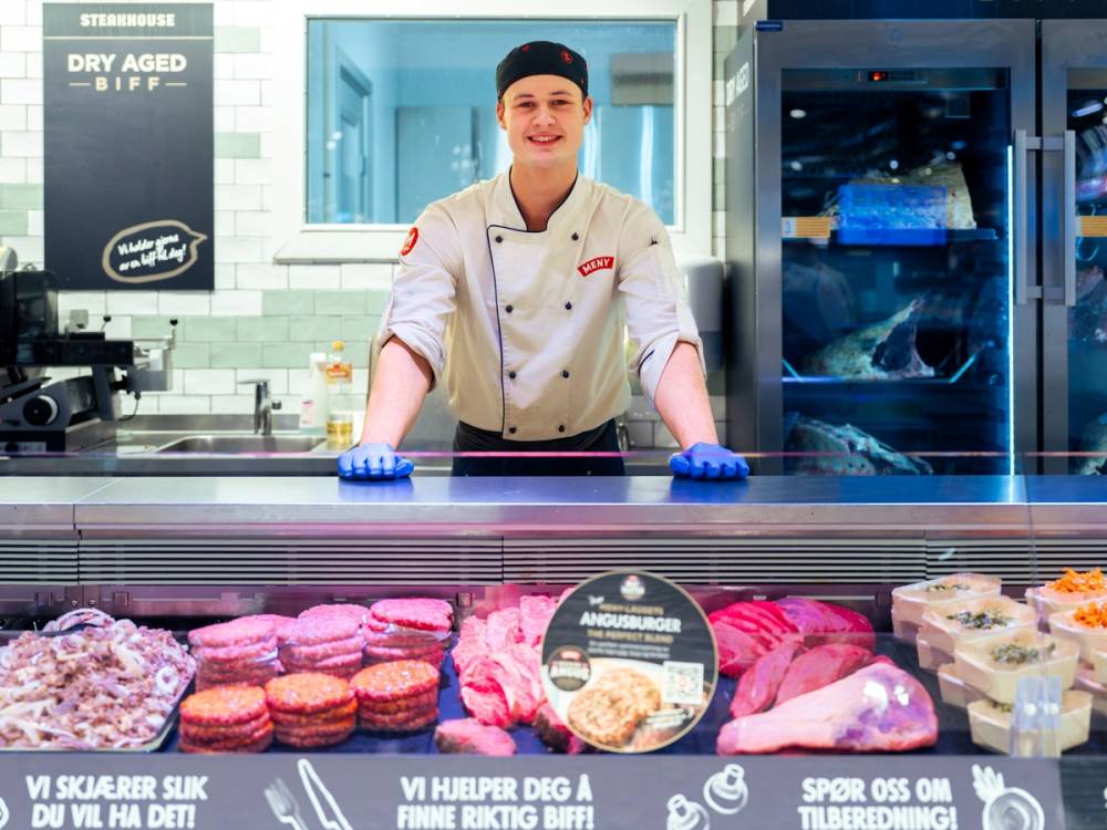En person i kokkeforkle med hvit uniform og blå hansker står bak en kjøtt- og bakevarer-stand i en butikk eller kafé, med kjøleskap i bakgrunnen.