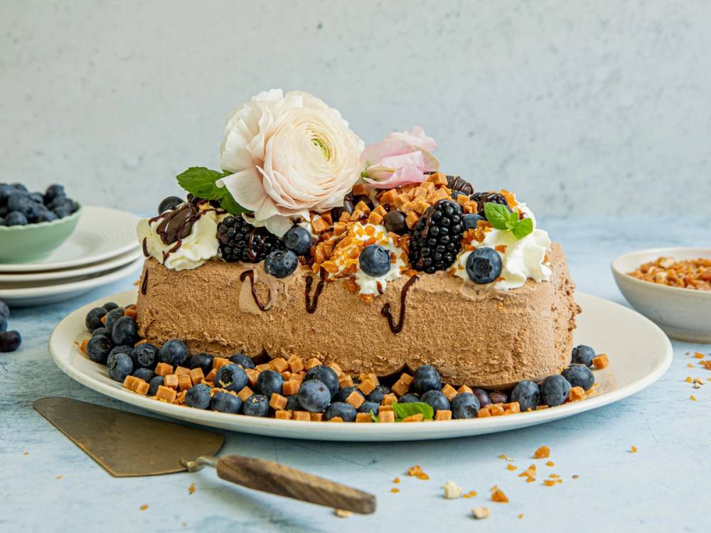 En iskremkake pyntet med bær, blomster og sjokoladesaus på en hvit tallerken, med en skje ved siden av.