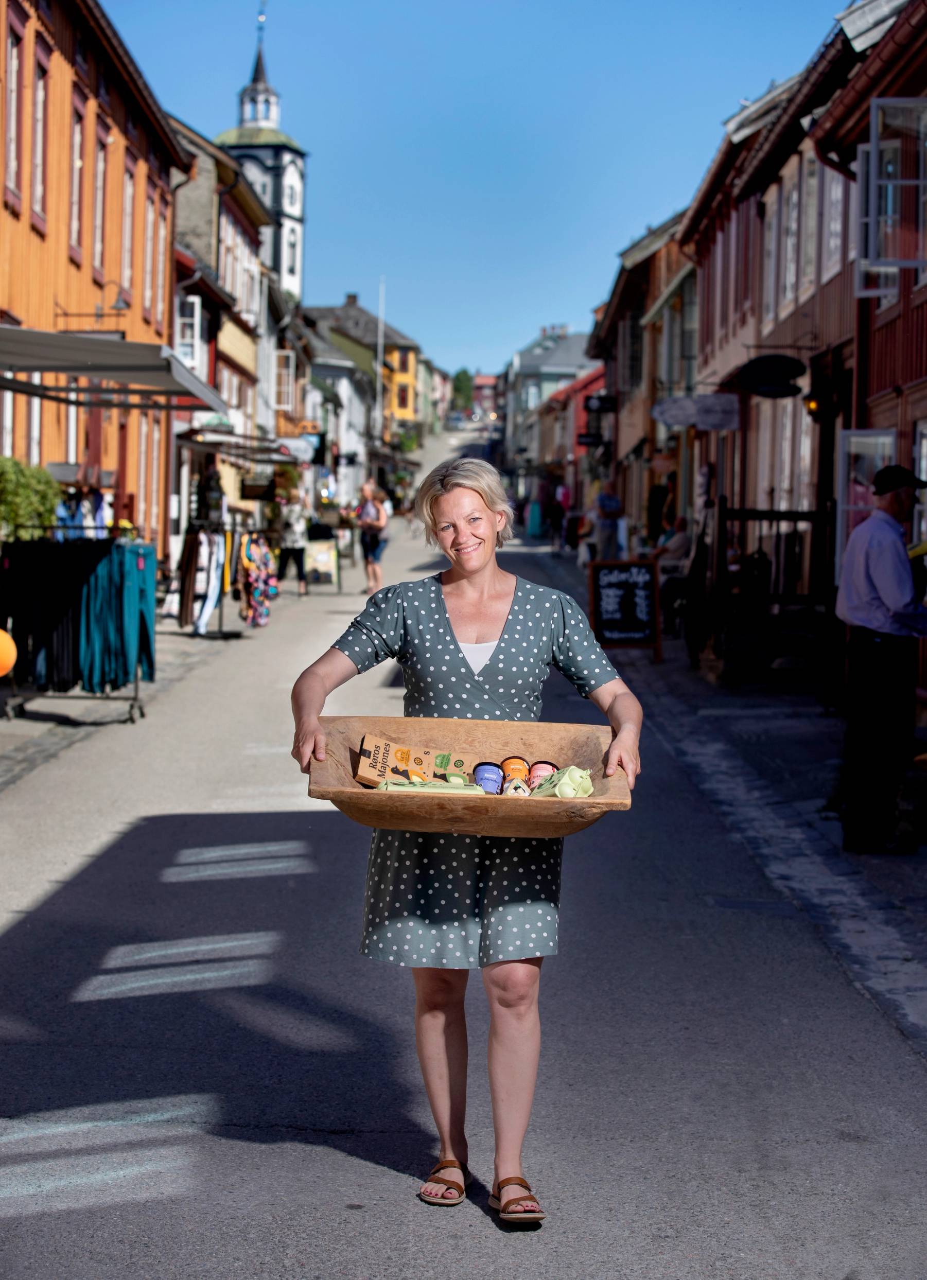 En person med kort hår og sommerkjole går i en gate med fargerike bygninger og bærer en kurv med småting.