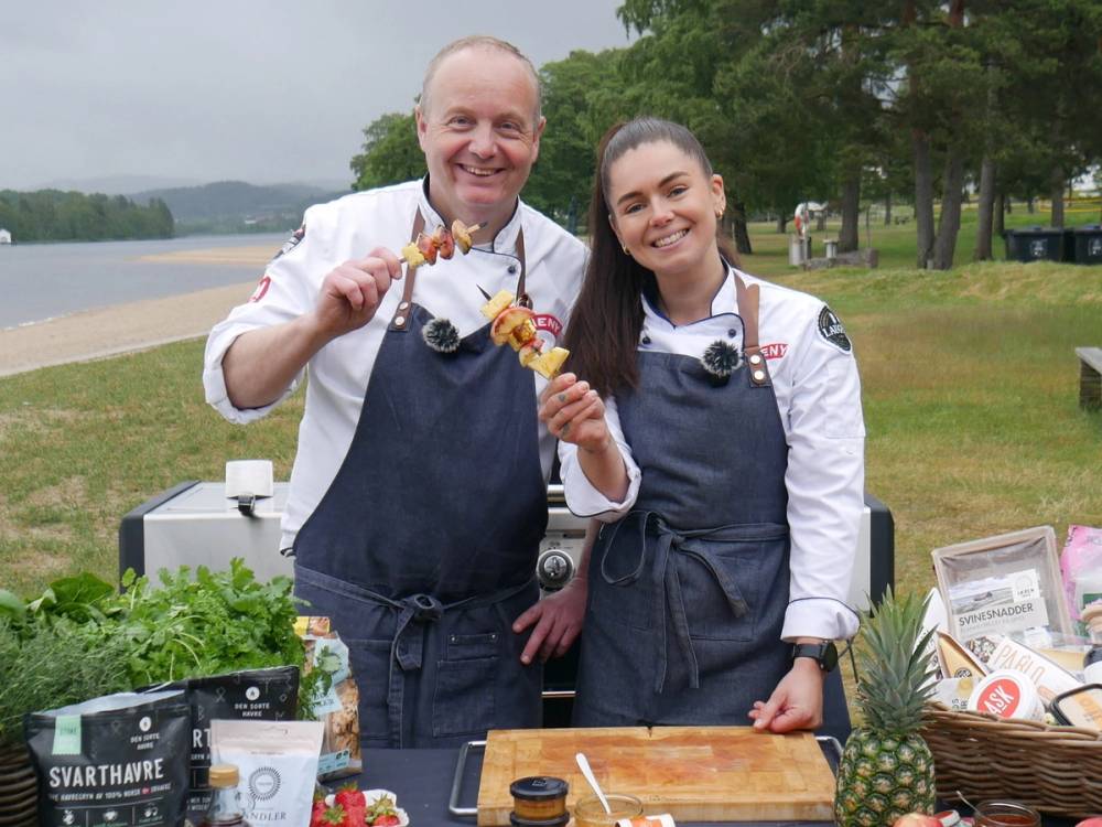 To personer med forkle som lager mat utendørs ved en grill, med grønt område og vann i bakgrunnen.