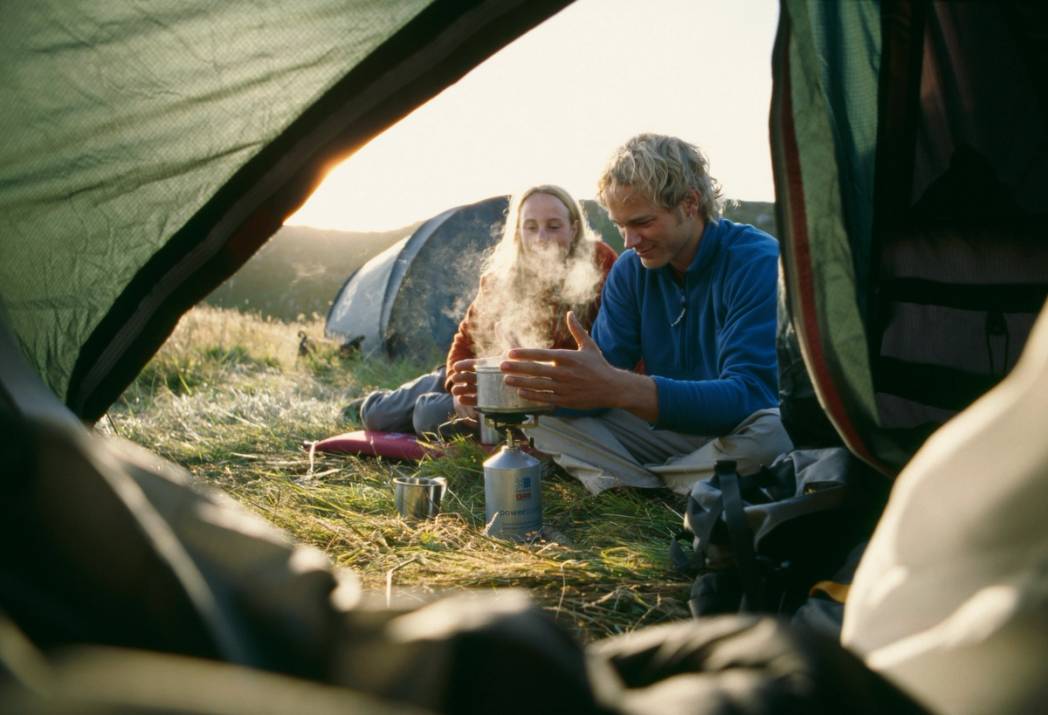 To personer sitter foran en campinghytte og nyter varme drikker i solskinnet, sett fra innsiden av en campingvogn.
