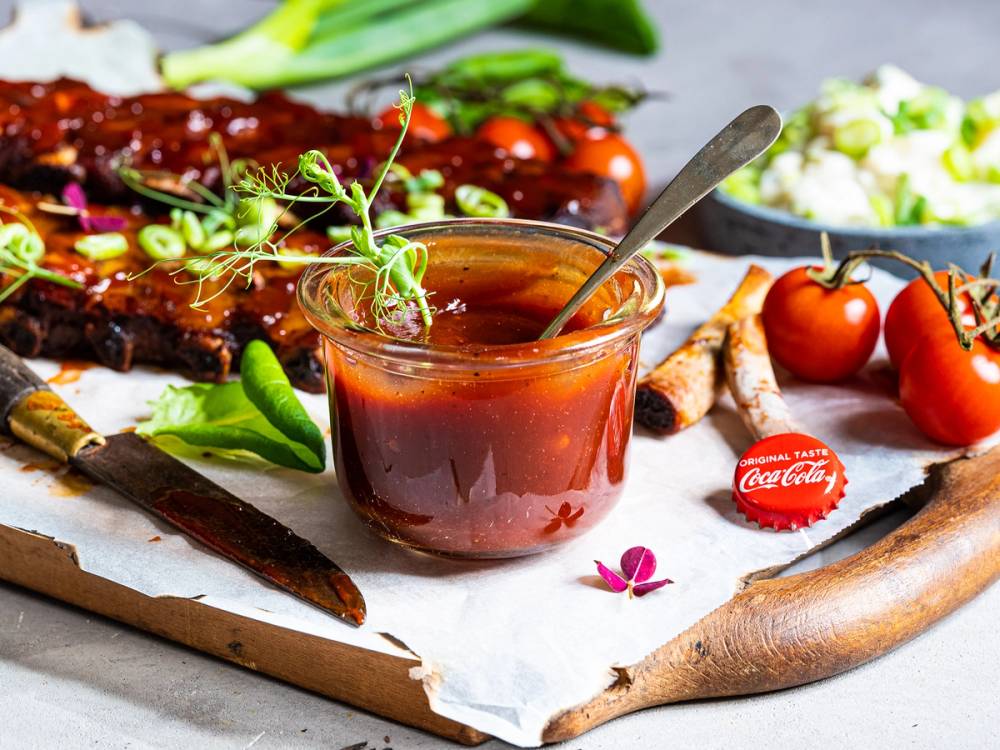 Marinade i en glassbolle med en skje, omgitt av grillet kjøtt, cherrytomater og grønnsaker på et serveringsbrett