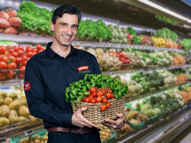 En ansatt står foran frukt- og grøntavdelingen og holder en kurv med tomater og grønne blader.