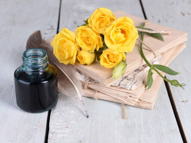 En bukett med gule roser ligger på en bunke av gamle aviser, med en svart inkwell og en fjærpenn ved siden av på et hvitt trebord.