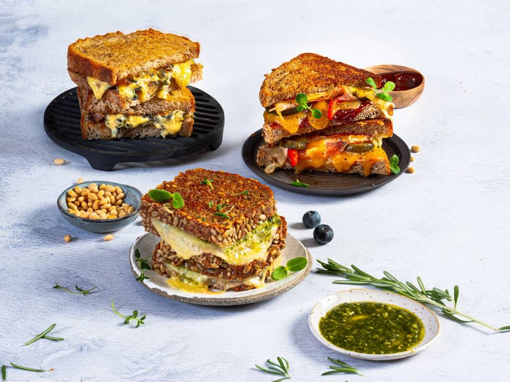 Tre ulike toast med pålegg og tilbehør på hvit bakgrunn, inkludert en liten skål med nøtter og en liten skål med grønn saus