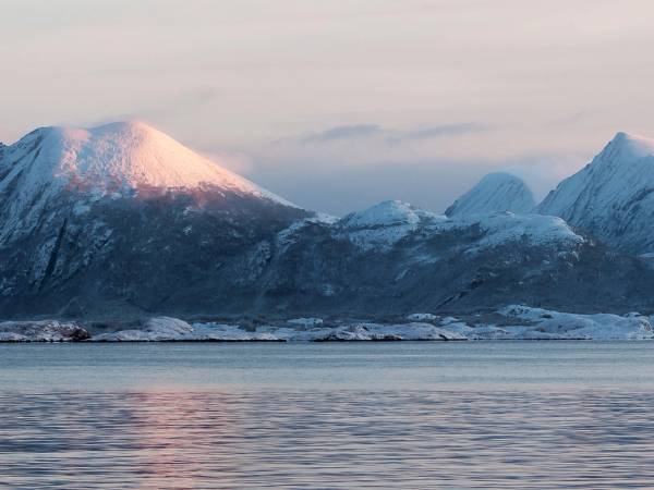 Fjell med snø på toppen bak en rolig fjord eller innsjø, med en overskyet himmel.