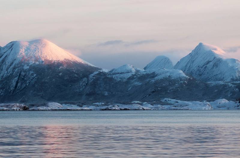 Fjell med snø på toppen bak en rolig fjord eller innsjø, med en overskyet himmel.
