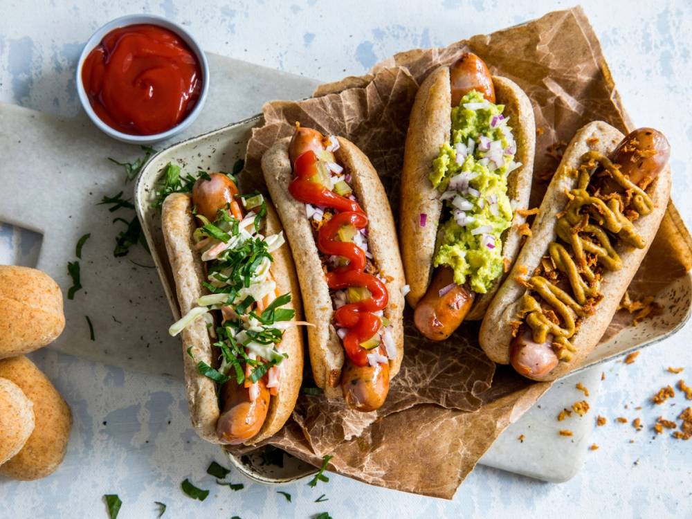 Tre pølser i brød med ulike tilbehør, servert på et papir underlag med en liten skål ketchup ved siden av.