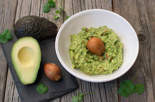 Avocado med stein i en skål, omgitt av halvert avocado og friske urter på en treplate