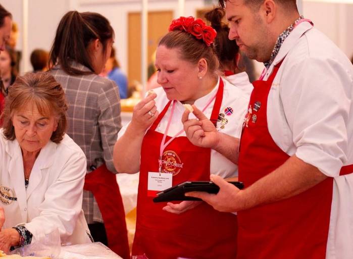 En gruppe mennesker, inkludert en kvinne med rød blomster i håret, ser på en enhet som en mann i hvit skjorte og rød forkle viser frem, i et innendørs arrangement.