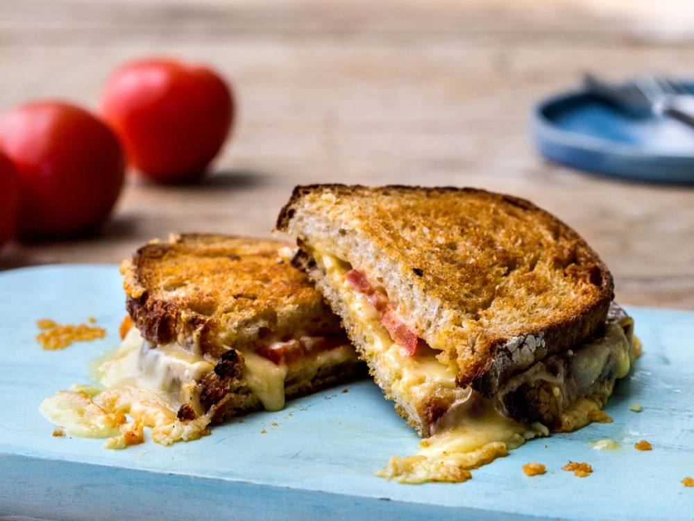 Et ostesmørbrød med norsk alpeost som er delt i to, på en blå tallerken med tomater i bakgrunnen.