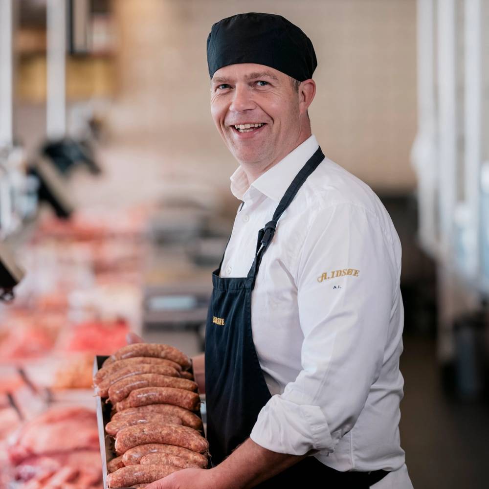 En person med hvit skjorte og svart forkle som står bak en kjøttdisk i en butikk eller på en restaurant, og holder opp en rekke kjøttdeigpakker.