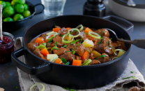 En gryte med gryterett som inneholder kjøtt, gulrøtter, poteter og løk, servert på et bord med grønnsaker og tilbehør.