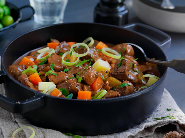 En gryte med gryterett som inneholder kjøtt, gulrøtter, poteter og løk, servert på et bord med grønnsaker og tilbehør.