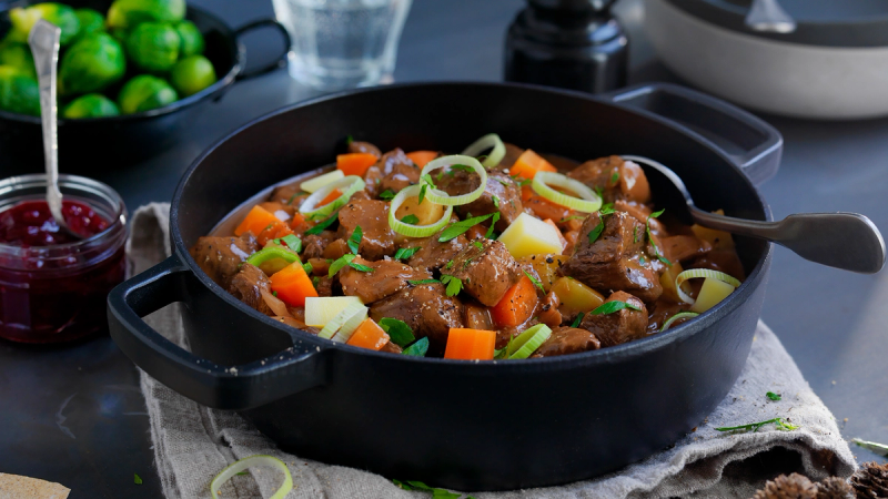 En gryte med gryterett som inneholder kjøtt, gulrøtter, poteter og løk, servert på et bord med grønnsaker og tilbehør.