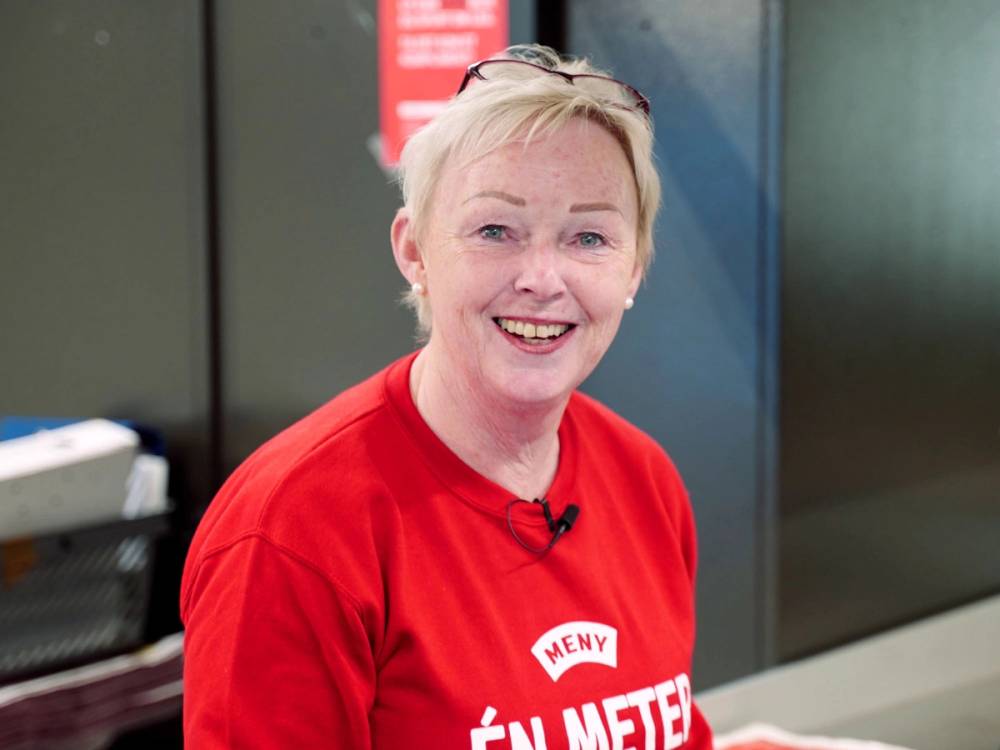 En person i rød MÅNY T-skjorte står i en butikk eller på et arbeidssted, med en vegg og utstyr i bakgrunnen.