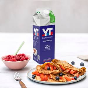 En flaske med yt-proteinmelk plassert ved siden av en tallerken med pannekaker toppet med frukt og bær, og en skål med bringebær.