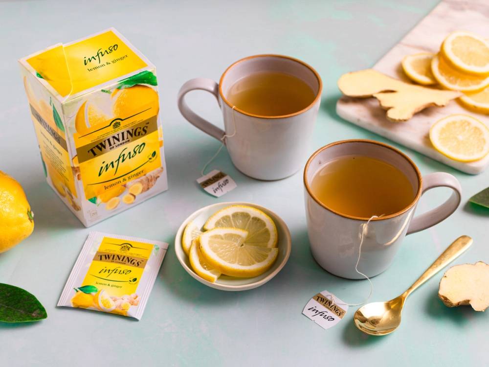 To kopper med te, en pakke Twinings Infuso sitron og ingefær, sitronbiter og ingefærskiver på et fat, og en skje på et lyst bord.