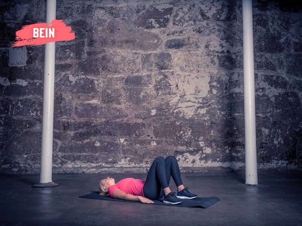 En kvinne ligger på ryggen på en treningsmatte og utfører seteløftøvelser i et treningsstudio med murvegg i bakgrunnen.