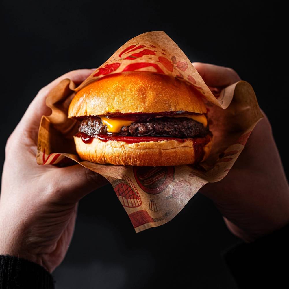 En person holder en burger med kjøtt, ost, og ketchup i et papirinnpakning mot en mørk bakgrunn.