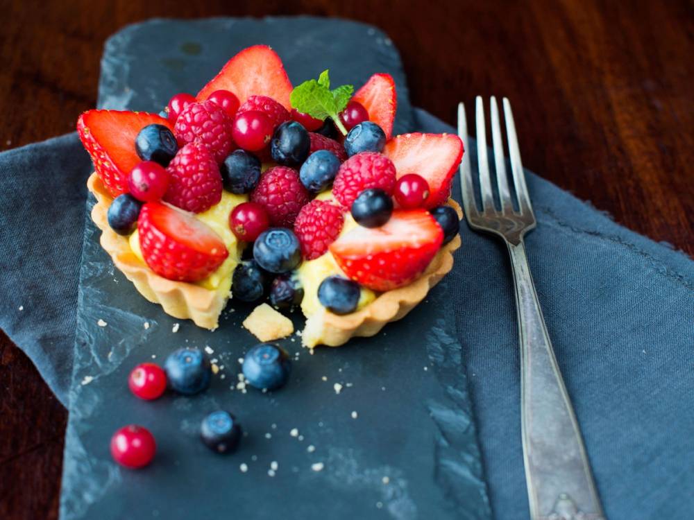 En fruktterte med jordbær, blåbær, rips og druer på en blå tallerken, med en gaffel ved siden av.