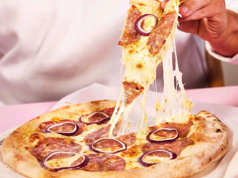 En person tar en bit av en pepperonipizza med ost som smelter og drar seg ut, på en rosa bordplate.