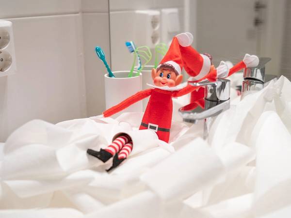 En liten julefigur med rød dress og hvitt skjegg står på en seng med hvite sengetøy, omgitt av tannbørster i kopper på badet.