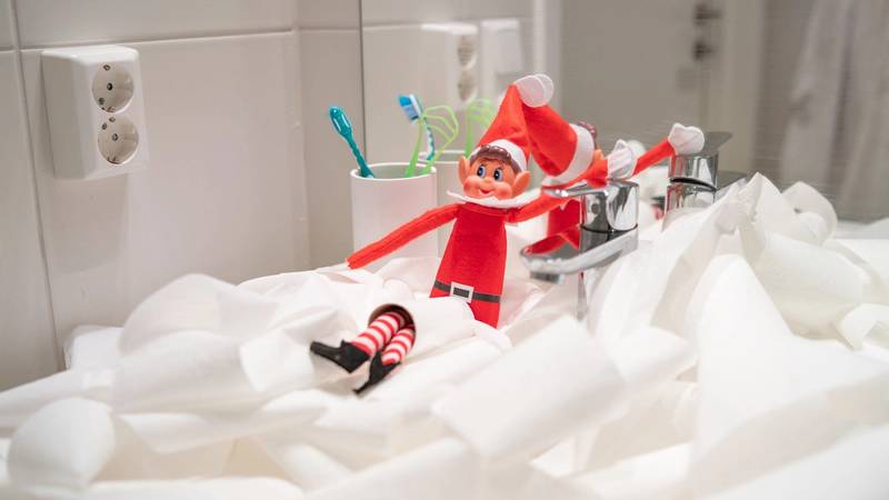 En liten julefigur med rød dress og hvitt skjegg står på en seng med hvite sengetøy, omgitt av tannbørster i kopper på badet.