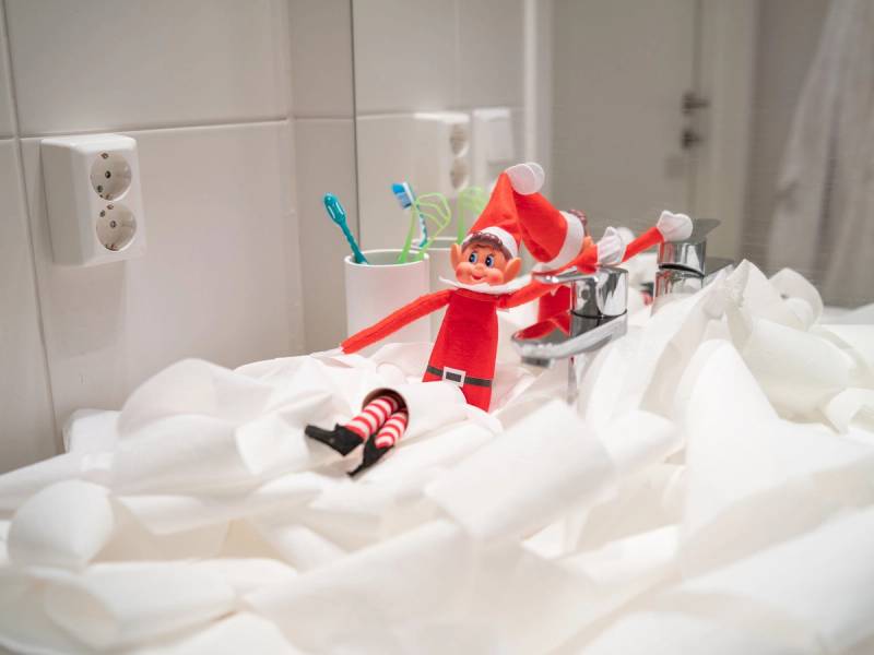 En liten julefigur med rød dress og hvitt skjegg står på en seng med hvite sengetøy, omgitt av tannbørster i kopper på badet.