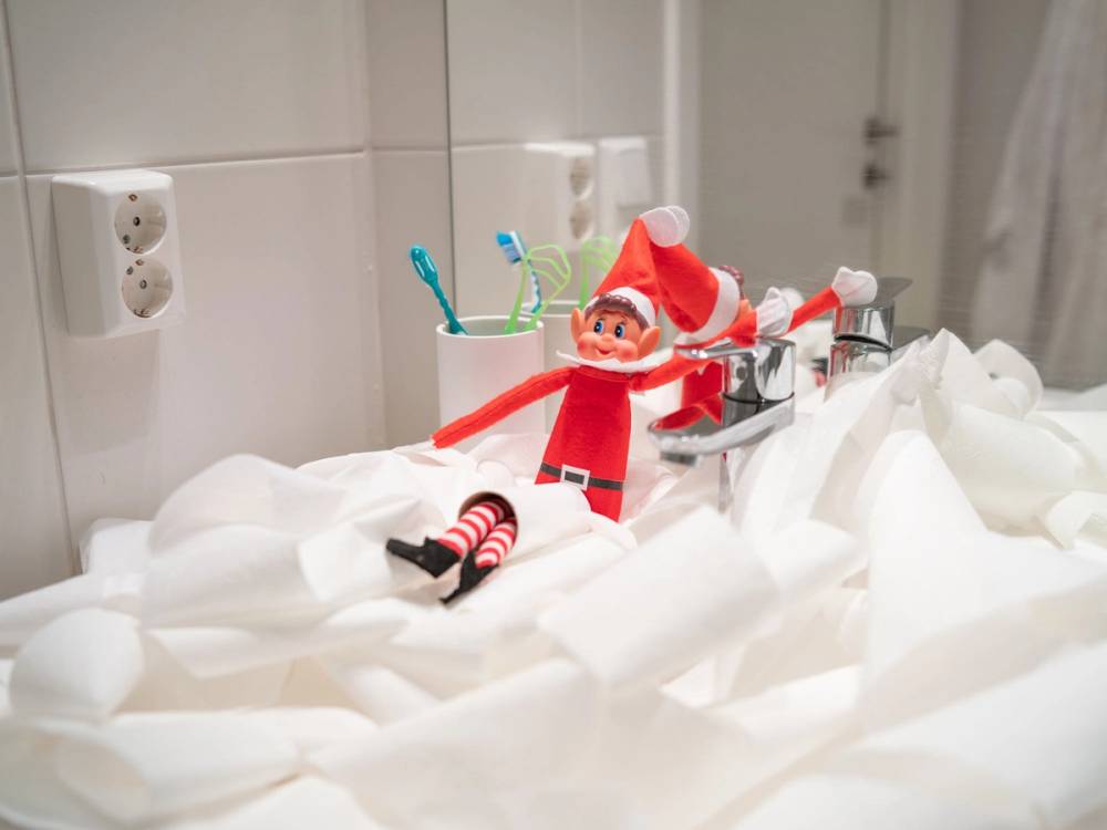 En liten julefigur med rød dress og hvitt skjegg står på en seng med hvite sengetøy, omgitt av tannbørster i kopper på badet.