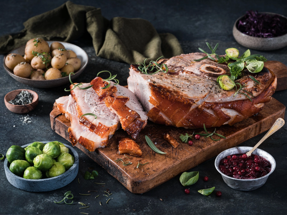 Skinkestek med en helstekt, gyllenbrun overflate, omgitt av små poteter, limebåter og bær, på et skjærebrett med urter og krydder rundt.