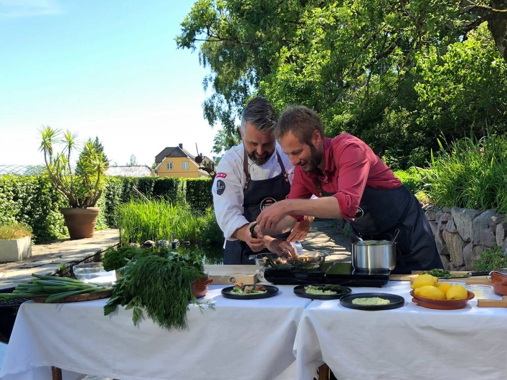 To personer lager mat utendørs på en hagefest, med grønnsaker og kokekar på et bord, omgitt av grønne trær og blomster.