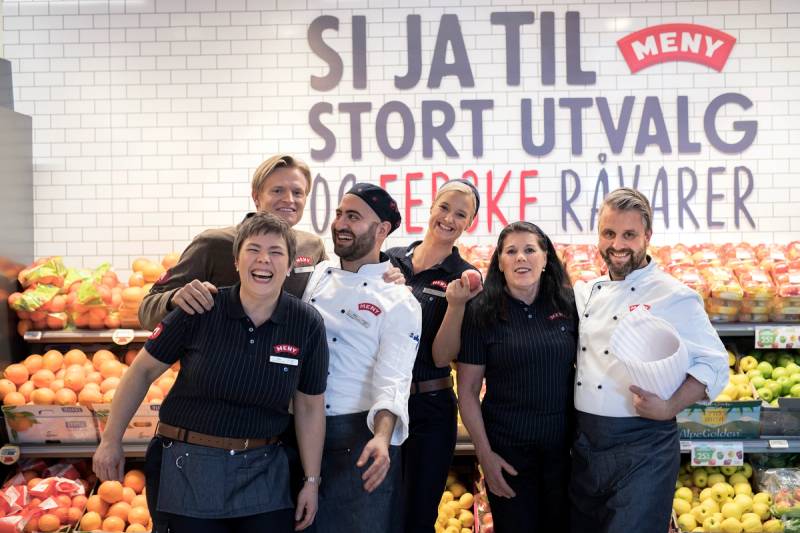 Gruppemedlemmer i en matbutikk står foran en vegg med frukt og grønnsaker, med en stor plakat som sier 'SI JA TIL STORT UTVALG' og 'MENY'.