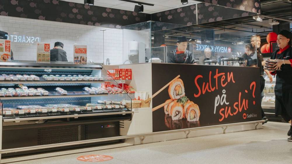 Sushireklame med teksten 'Sulten på sushi?' på en butikkdisk, med ansatte og kunder i bakgrunnen.