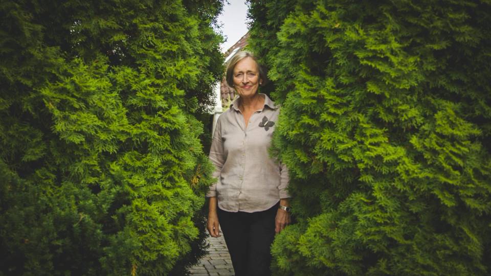 En person står mellom grønne busker på en sti utendørs, med en bygning i bakgrunnen.