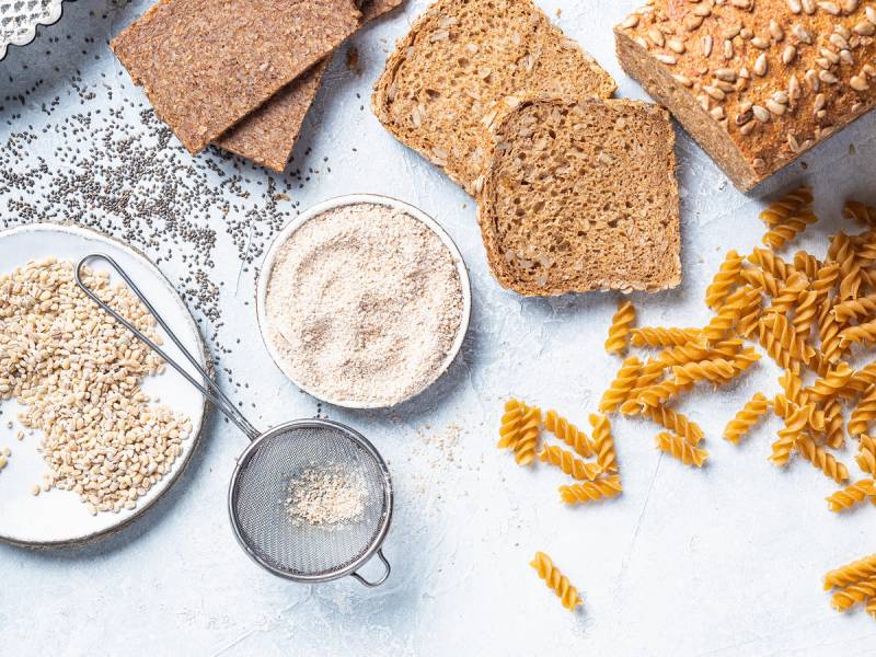 Grov fullkornsbrød, pasta og mel i ulike beholdere på et kjøkkenbord.