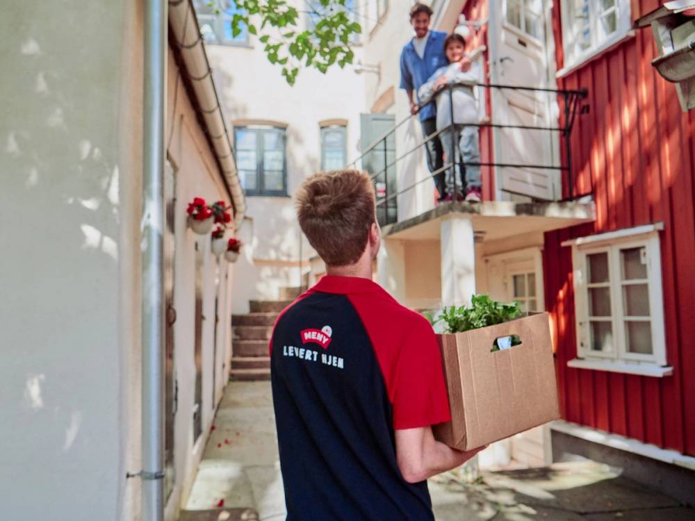 En mann i en Meny-levert hjem-uniform leverer en pakke til to personer på en balkong i en boligblokk.