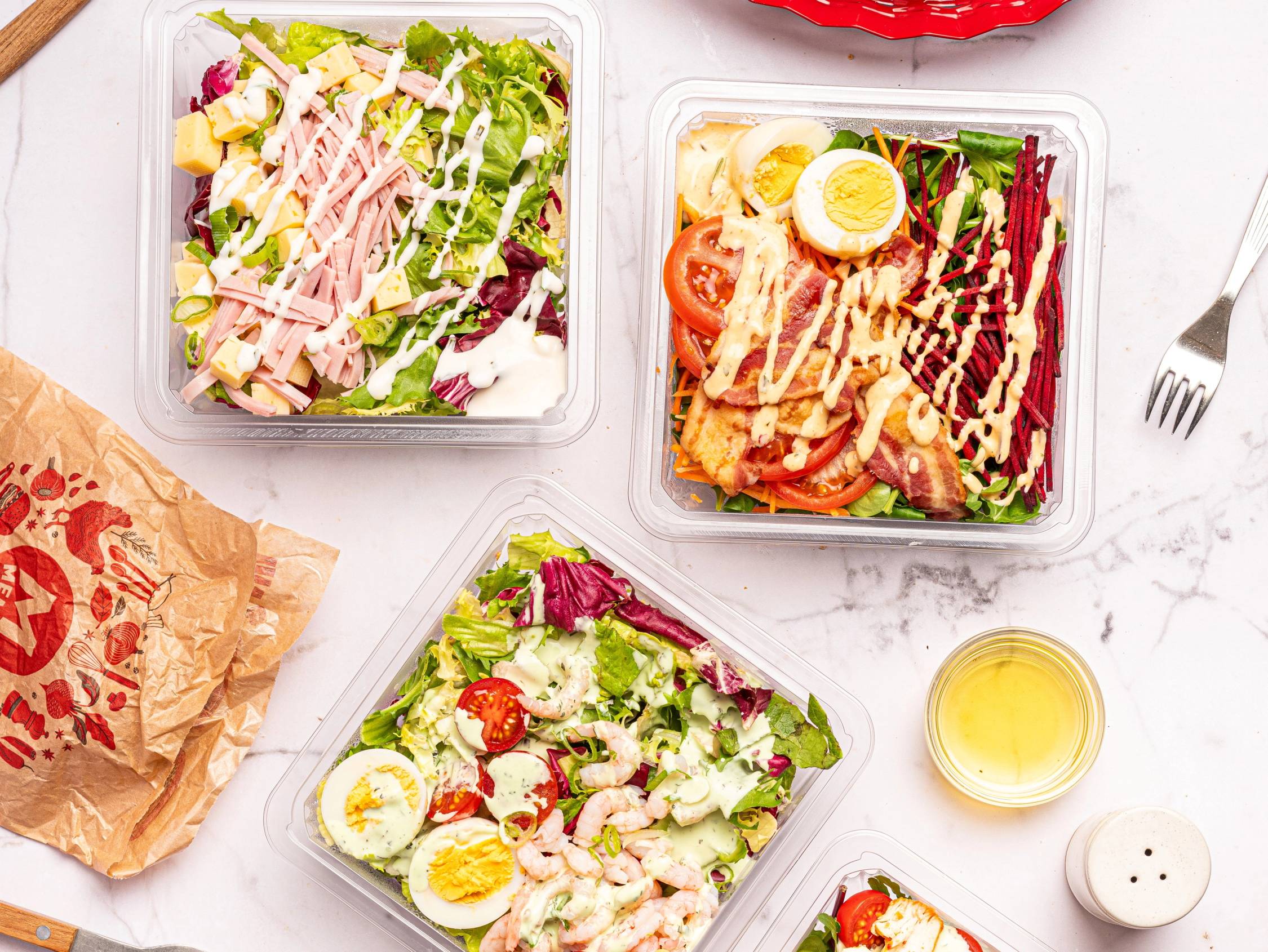 Tre ferdigpakkede salatbokser med ulike salattyper på et hvitt bord, med en liten bolle med dressing og en gaffel i nærheten.