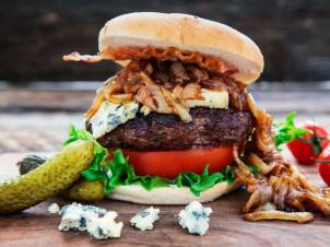 En burger med blåskimmelost, salat, tomat og karamellisert løk i et hamburgerbrød, med tilbehør av sylteagurk og blåmuggost på siden.