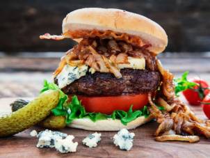 En burger med blåskimmelost, salat, tomat og karamellisert løk i et hamburgerbrød, med tilbehør av sylteagurk og blåmuggost på siden.