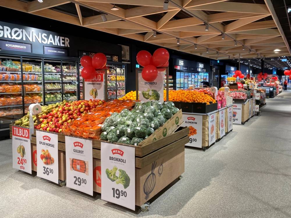 Frukt- og grønnsaksavdeling i en dagligvarebutikk med pappesker fylt med appelsiner, brokkoli og andre grønnsaker, dekorert med røde ballonger.
