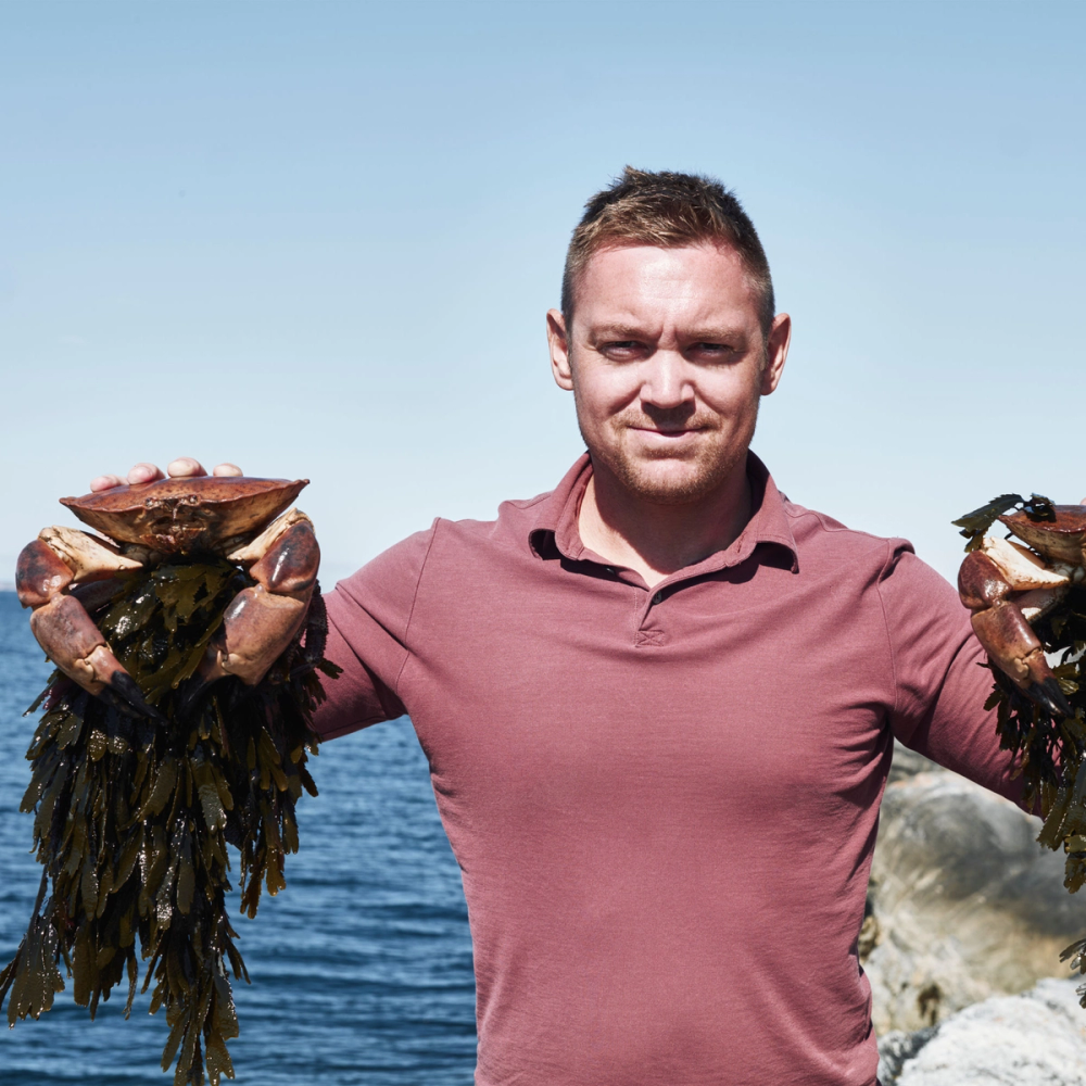 En person holder opp to store hummerkreps foran en sjø med steiner og blå himmel i bakgrunnen.