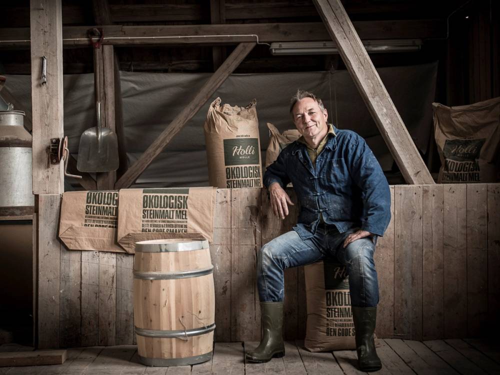 En person i jeans og jakke sitter på en trebenk i et rom med sekker og verktøy, og det er en barrel foran personen.