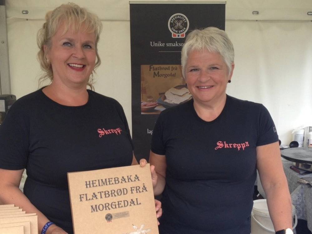 To personer står sammen bak en bord med en plakat som sier 'HEIMEBAKA FLATBRØD FRA MORGENDAL', og de har på seg svarte t-skjorter med 'Shrepp' på, i en innendørs setting.