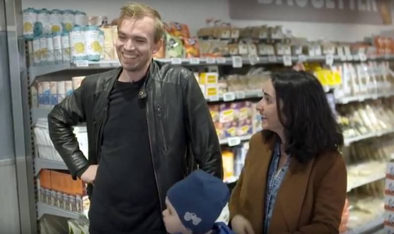 To personer snakker i en dagligvarebutikk med hyller fylt med matvarer i bakgrunnen.