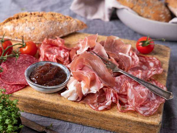Frokostbord med spekemat, tomater, brød og syltetøy på et trefat.